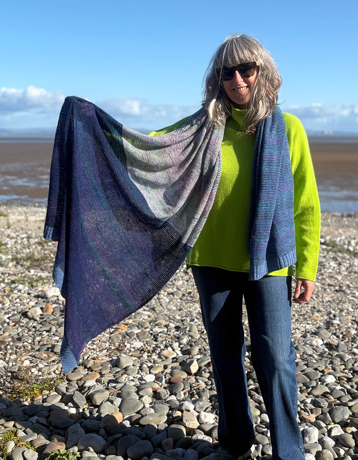 A woman standing on a rocky beach holds up a large square shawl with a variegated color pattern of purple, blue, gray, and green.