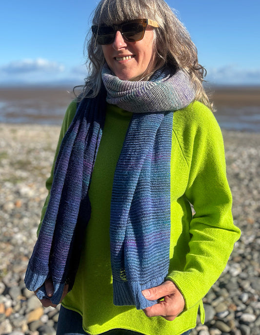 A woman wearing a bright lime green sweater, dark sunglasses, and a thick knitted scarf with blue, purple, and gray stripes, standing outdoors on a pebble beach with water and sky in the background.