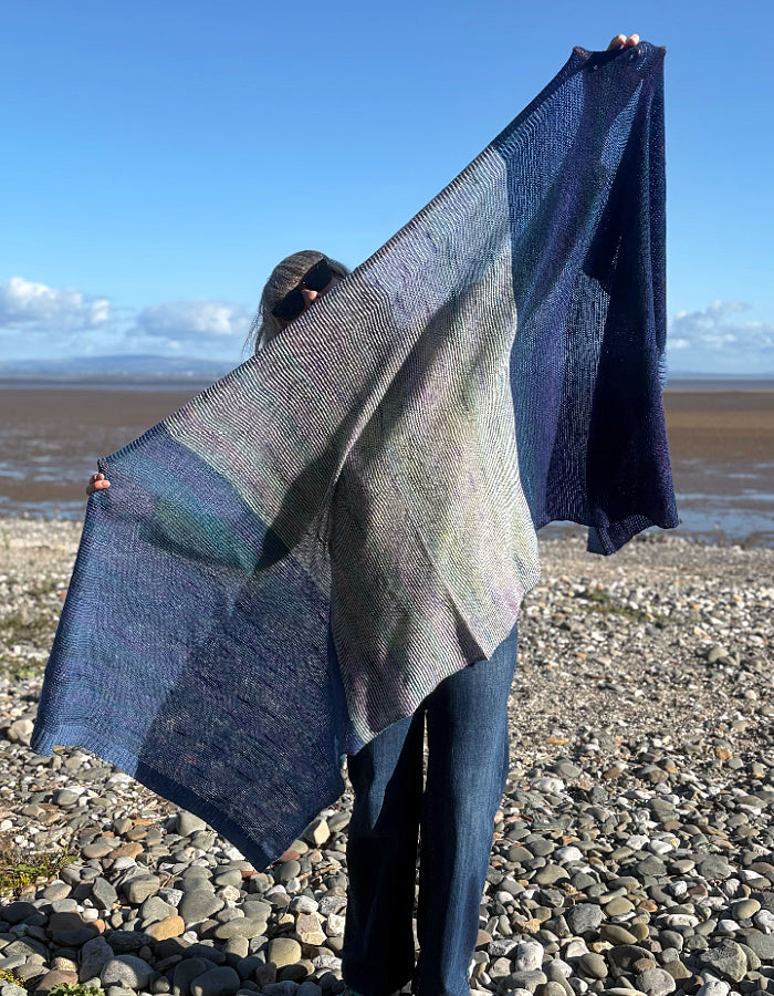 A person standing on a pebble beach holds up a large open-knit shawl with a blue, gray, and muted green color-blocked pattern against a background of sky and water.