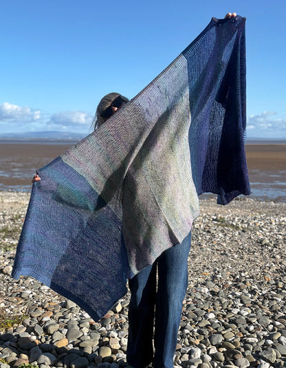 A person standing on a pebble beach holds up a large open-knit shawl with a blue, gray, and muted green color-blocked pattern against a background of sky and water.
