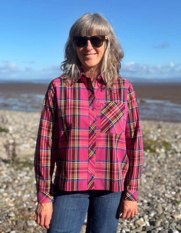 A woman wearing a pink and red plaid button-up shirt with yellow and black stripes, standing on a rocky beach with water and a blue sky in the background.