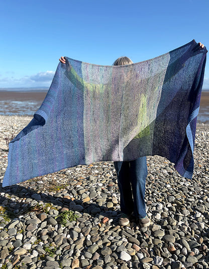 A person standing on a pebble beach holds up a large knitted shawl with vertical color-blocked panels in blue, purple, and green tones under a clear blue sky.