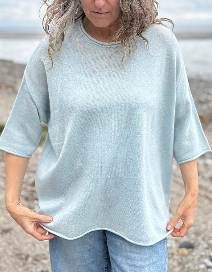 Woman wearing a light blue cashmere jumper standing on a beach with ocean and sky in the background