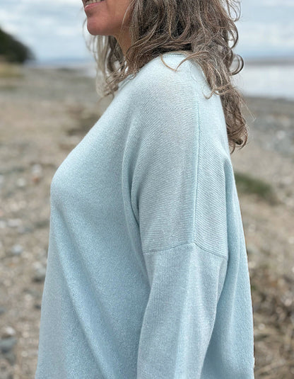 Woman wearing a light blue cashmere jumper standing on a beach with ocean and sky in the background