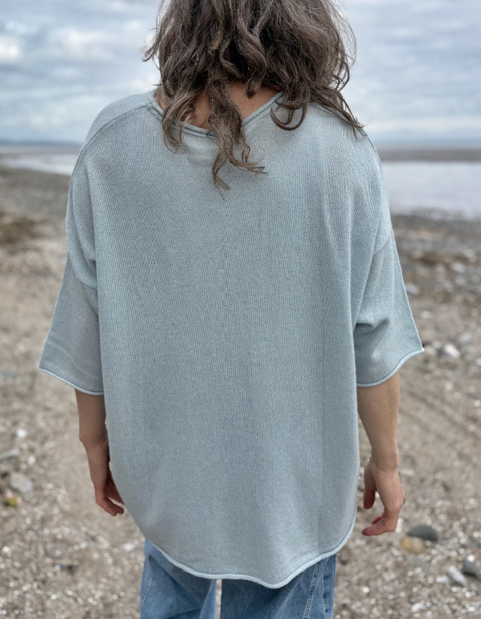 Woman wearing a light blue cashmere jumper standing on a beach with ocean and sky in the background