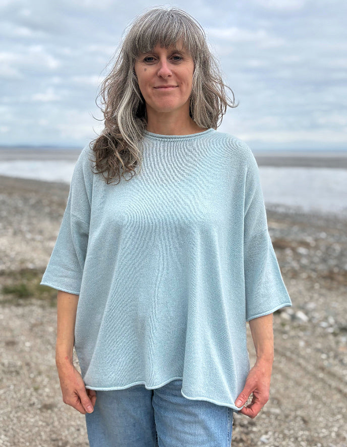 Woman wearing a light blue cashmere jumper standing on a beach with ocean and sky in the background