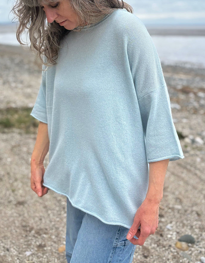 Woman wearing a light blue cashmere jumper standing on a beach with ocean and sky in the background