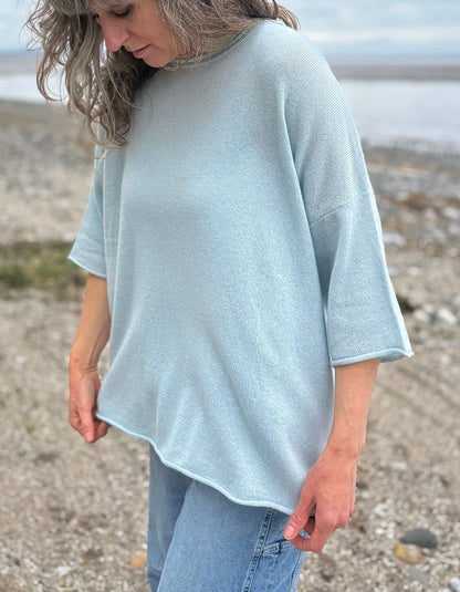 Woman wearing a light blue cashmere jumper standing on a beach with ocean and sky in the background
