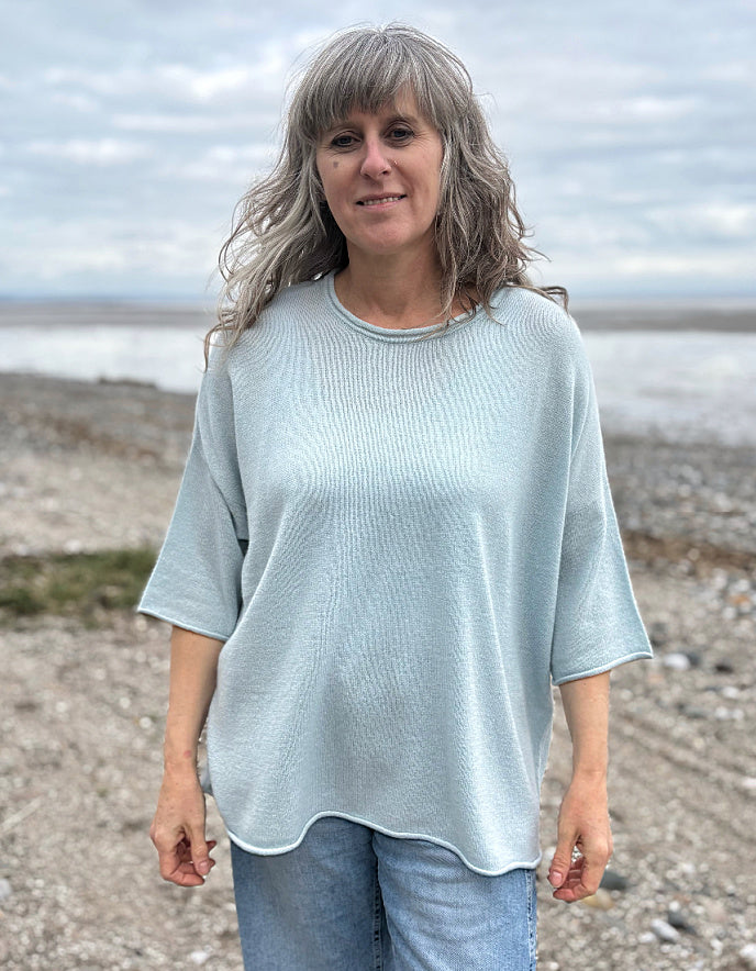 Woman wearing a light blue cashmere jumper standing on a beach with ocean and sky in the background