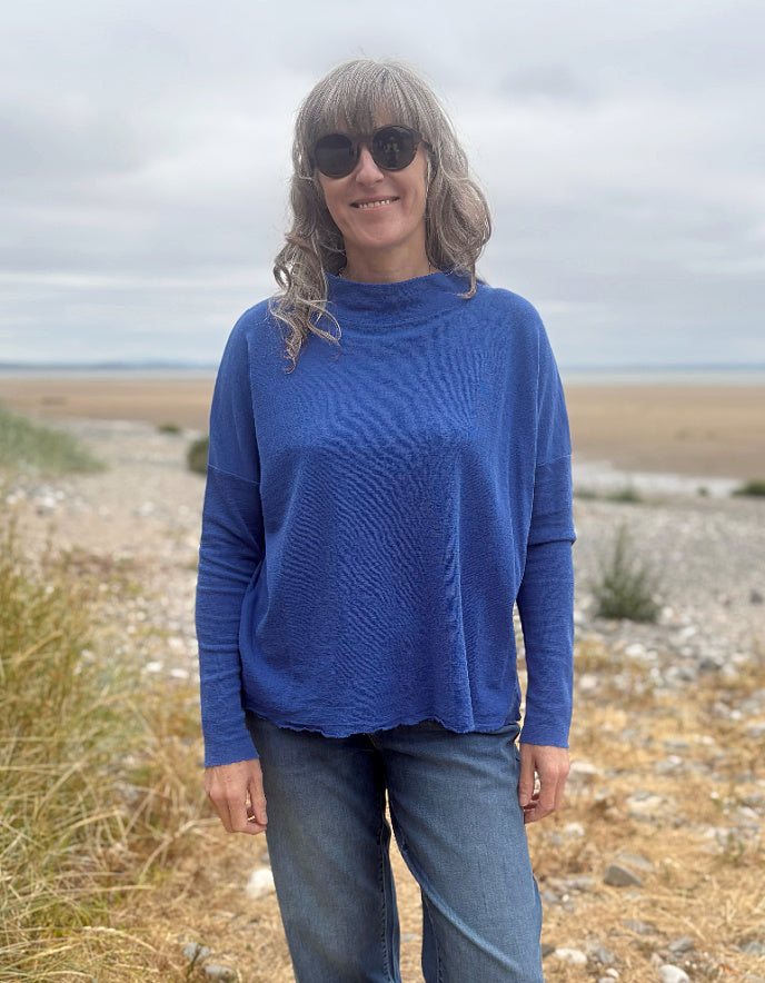 A woman wearing a textured royal blue long-sleeve knit top and blue jeans stands outdoors on a rocky, grassy shoreline under an overcast sky.