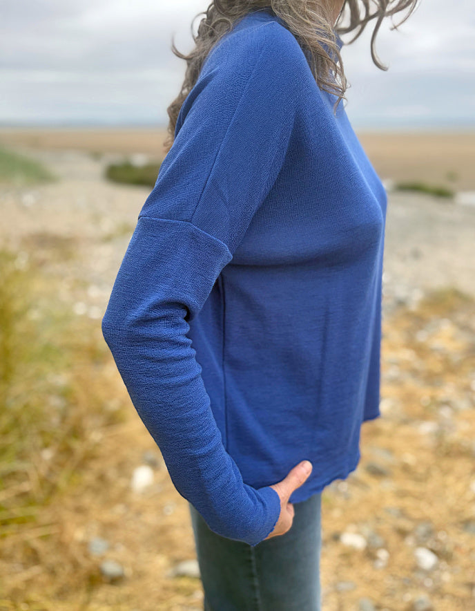 A woman wearing a blue long-sleeve knit sweater, shown from the side in an outdoor coastal setting with a blurred beach and grassy background.