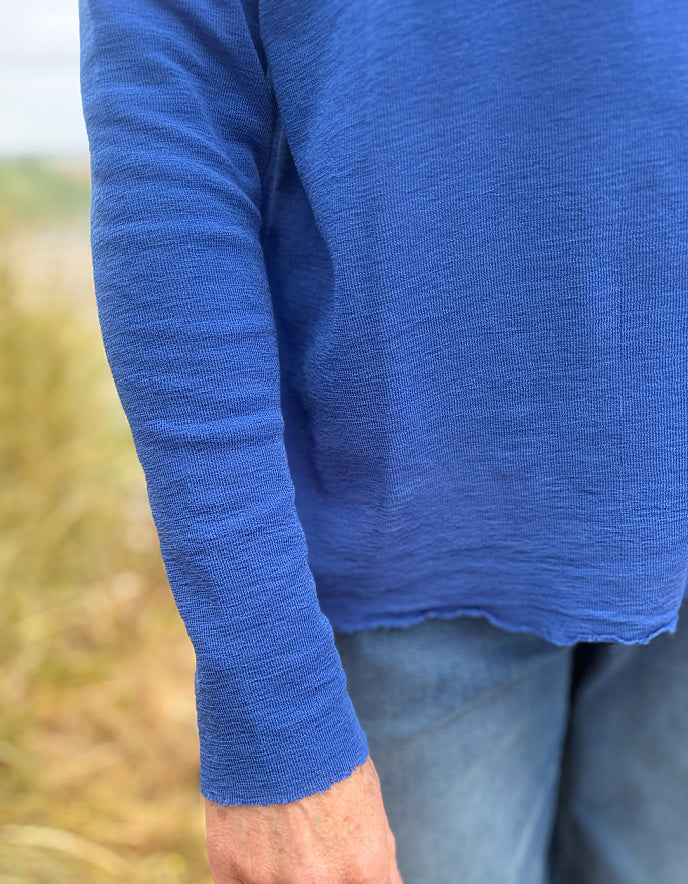 A close-up of a person wearing a textured royal blue long-sleeve top and light blue jeans, standing outdoors with a blurred natural background.