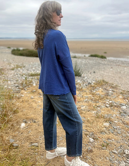 A woman wearing a blue knit long-sleeve top, dark blue jeans, and white sneakers stands on a rocky beach path with a sandy shoreline and cloudy sky in the background.