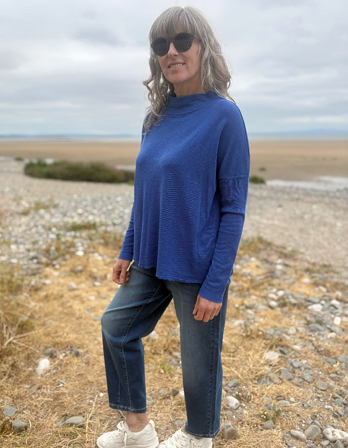 A woman wearing a textured royal blue long-sleeve mock neck top, dark blue jeans, and white sneakers, standing outdoors on a rocky, dry beach landscape under an overcast sky.