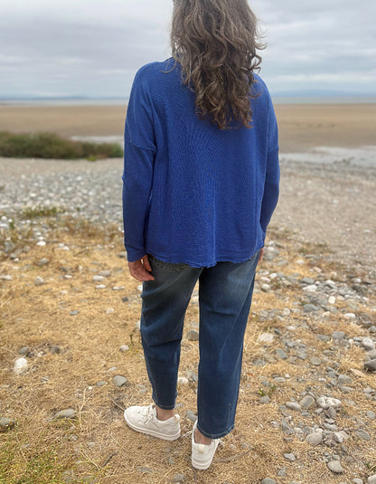 A person standing outdoors on a rocky, dry shoreline wearing a blue ribbed knit sweater, dark blue jeans, and white sneakers, photographed from behind.