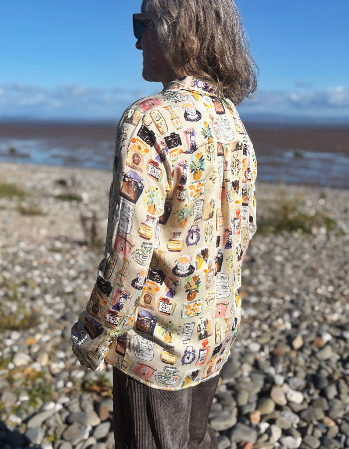 A woman with gray hair and sunglasses stands on a rocky beach wearing a beige button-up silky shirt covered in colourful illustrated print and dark brown cord wide leg pants, with water and blue sky in the background.