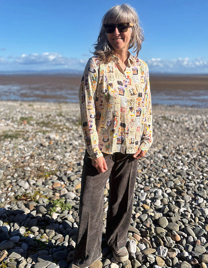 A woman with gray hair and sunglasses stands on a rocky beach wearing a beige button-up silky shirt covered in colourful illustrated print and dark brown cord wide leg pants, with water and blue sky in the background.