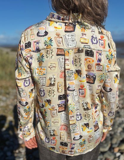 A woman with gray hair and sunglasses stands on a rocky beach wearing a beige button-up silky shirt covered in colourful illustrated print and dark brown cord wide leg pants, with water and blue sky in the background.