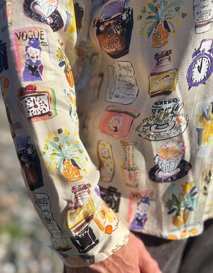 A close-up of a person wearing a cream-colored long-sleeve shirt with a colorful, whimsical illustration pattern featuring teacups, flowers, and vintage objects.