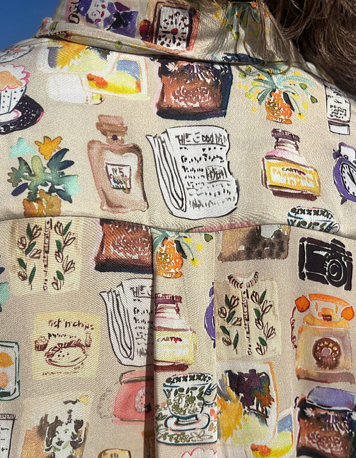 A close-up of a beige fabric garment featuring a whimsical, illustrated collage print of perfume bottles, notebooks, plants, and vintage objects in a watercolor style.