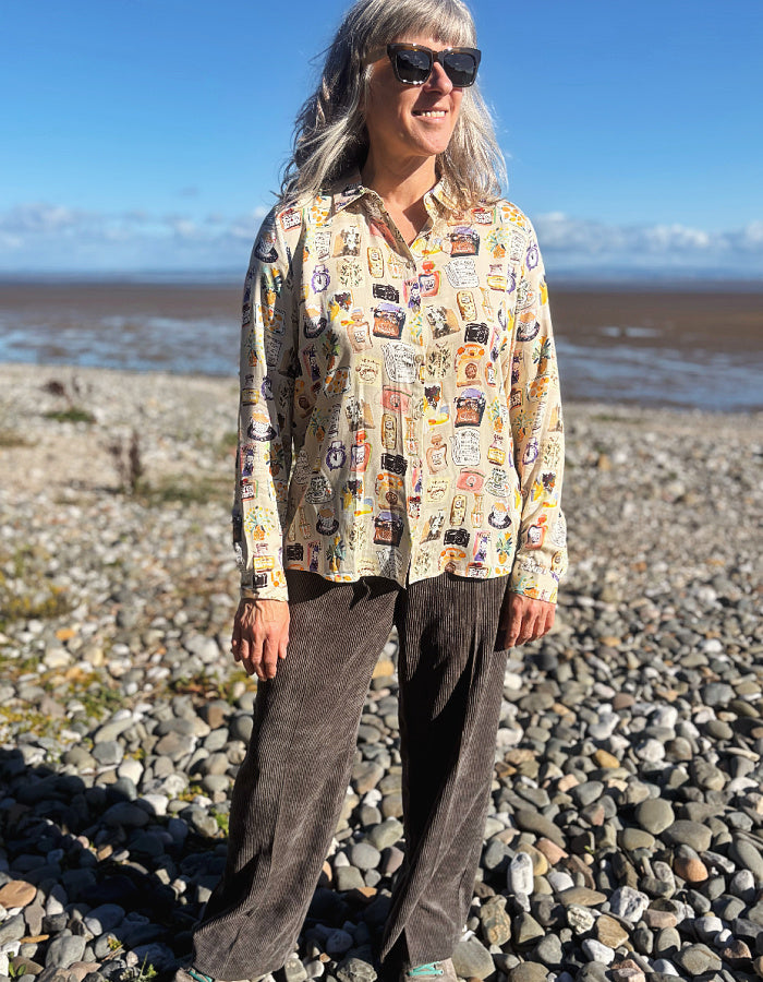 A woman with gray hair and sunglasses stands on a rocky beach wearing a beige button-up silky shirt covered in colourful illustrated print and dark brown cord wide leg pants, with water and blue sky in the background.