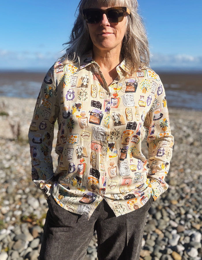 A woman with gray hair and sunglasses stands on a rocky beach wearing a beige button-up silky shirt covered in colourful illustrated print and dark brown cord wide leg pants, with water and blue sky in the background.