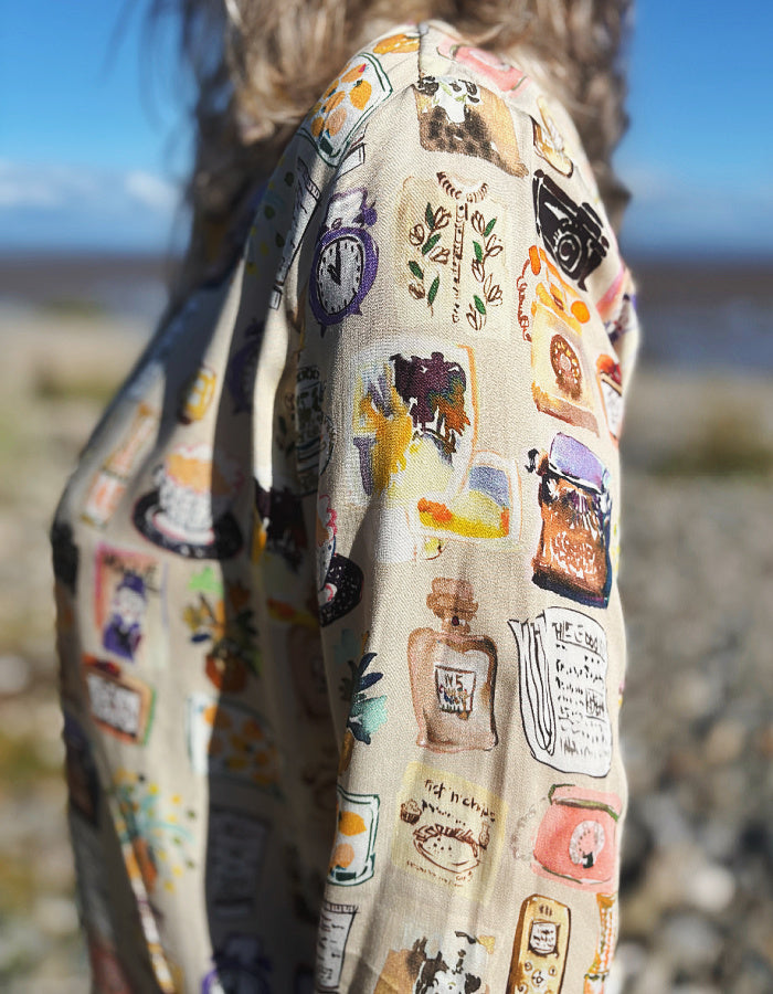 A woman with gray hair and sunglasses stands on a rocky beach wearing a beige button-up silky shirt covered in colourful illustrated print and dark brown cord wide leg pants, with water and blue sky in the background.