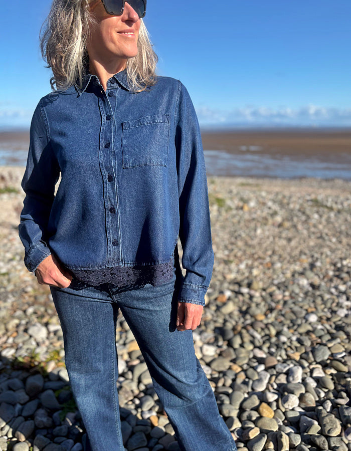 A woman wearing a blue denim long-sleeve button-up shirt and matching blue jeans, standing on a pebble beach with water and sky in the background.