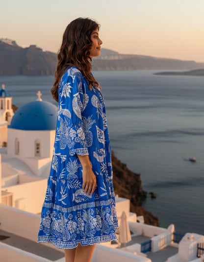 Woman in a blue dress with white patterns standing in front of a scenic view with mountains and water.