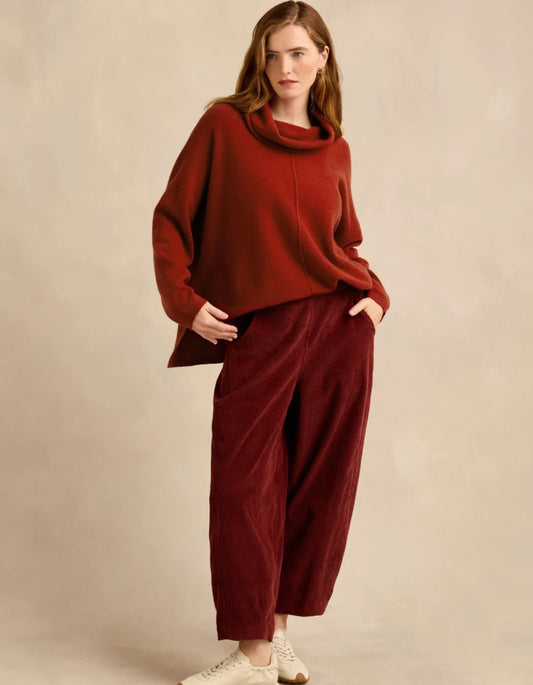 A woman modeling a rust red cowl-neck knit sweater and matching deep burgundy corduroy wide-leg pants against a warm beige studio backdrop.