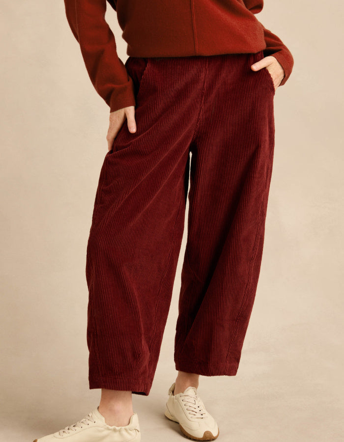 A person wearing loose, wide-leg corduroy pants in a deep burgundy red, paired with a matching rust-brown top and off-white sneakers, standing against a plain beige studio background.