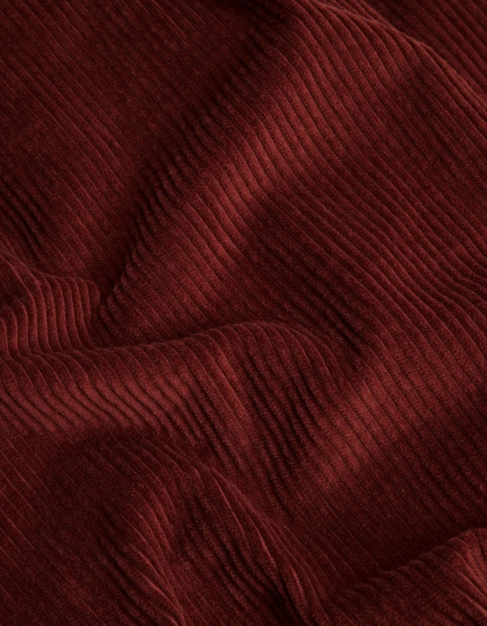 A close-up of draped corduroy fabric in a deep burgundy red color, showing the ribbed texture and soft folds.