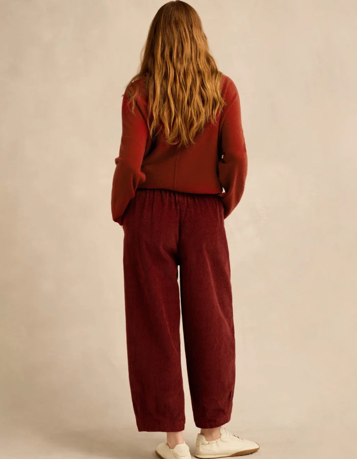 A woman viewed from behind wearing a rust-red long-sleeve top and matching dark red wide-leg pants, paired with white sneakers, against a neutral beige studio backdrop.