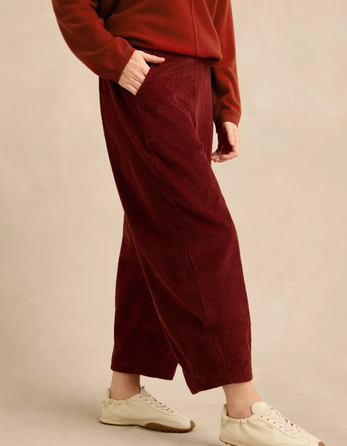 A person wearing wide-leg burgundy corduroy pants, a rust-colored long-sleeve top, and cream sneakers, standing against a neutral beige studio backdrop.