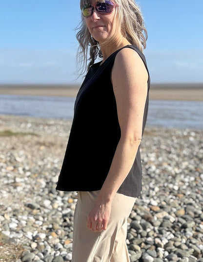 Woman standing on a pebbly beach wearing a black sleeveless top and sunglasses.