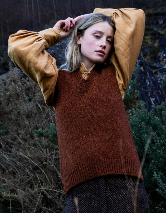 Woman wearing a copper brown donegal tweed tank top with a yellow shirt underneath, standing in a natural setting.