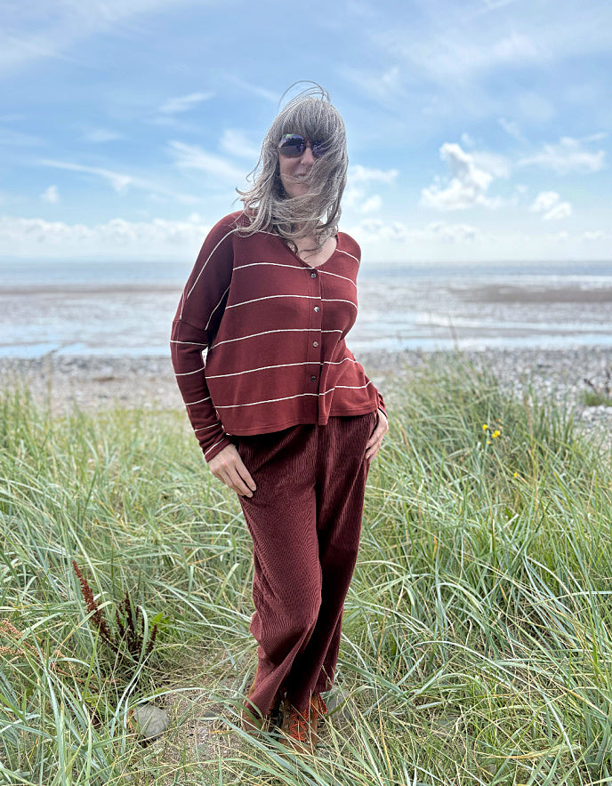 Person wearing a copper striped cardigan and pants standing in a grassy field with a beach in the background.
