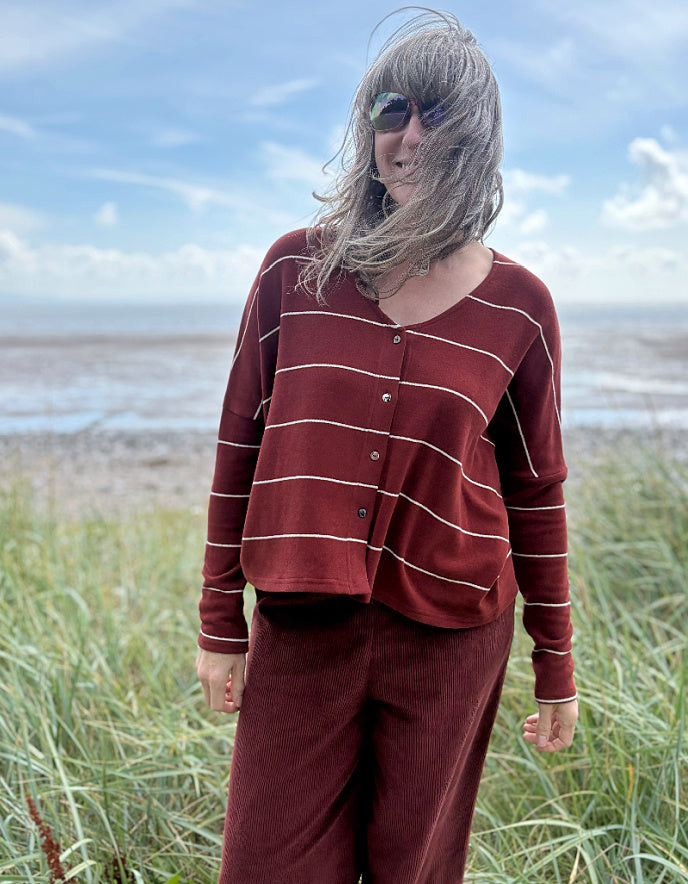 Person wearing a copper striped cardigan and pants standing in a grassy field with a beach in the background.