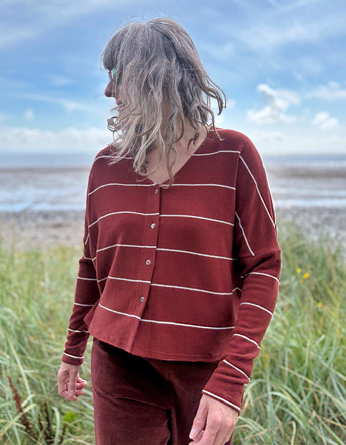 Person wearing a copper striped cardigan and pants standing in a grassy field with a beach in the background.
