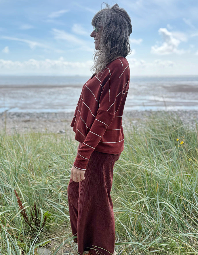 Person wearing a copper striped cardigan and pants standing in a grassy field with a beach in the background.
