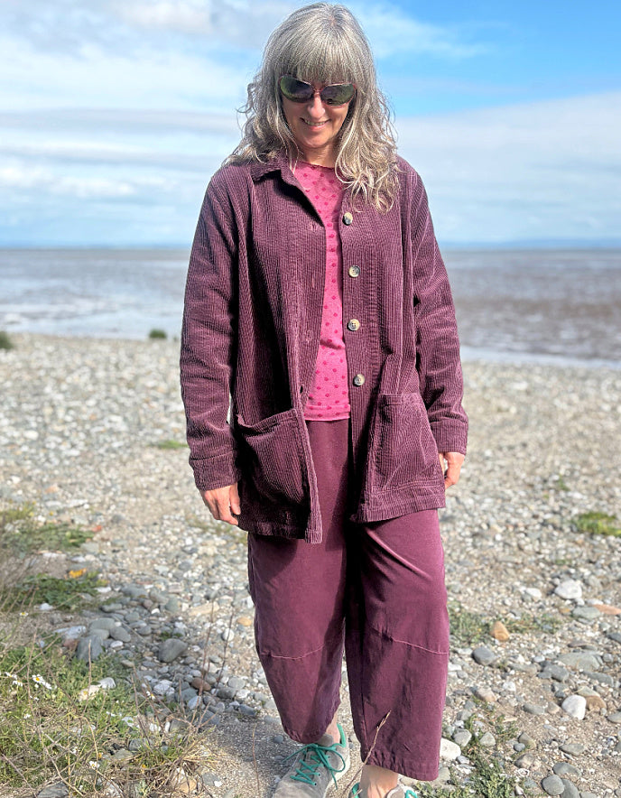Woman standing on a beach wearing a purple jumbo corduroy blazer and pink shirt.