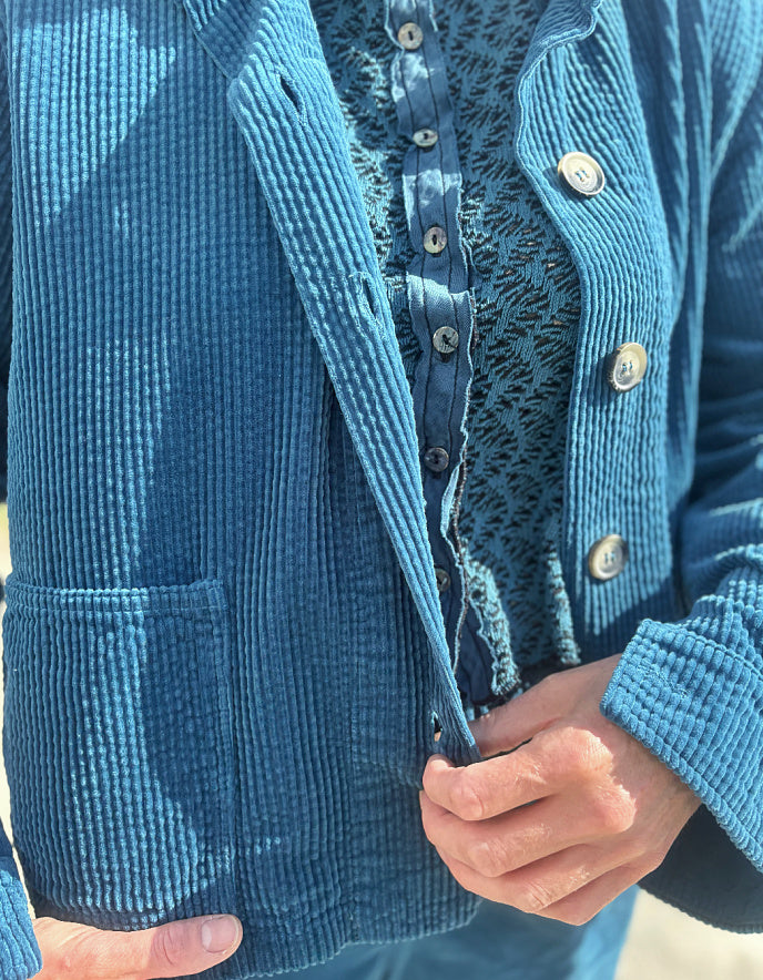 A close-up of a person wearing a textured blue corduroy jacket over a patterned blue top, with hands adjusting the front of the jacket.