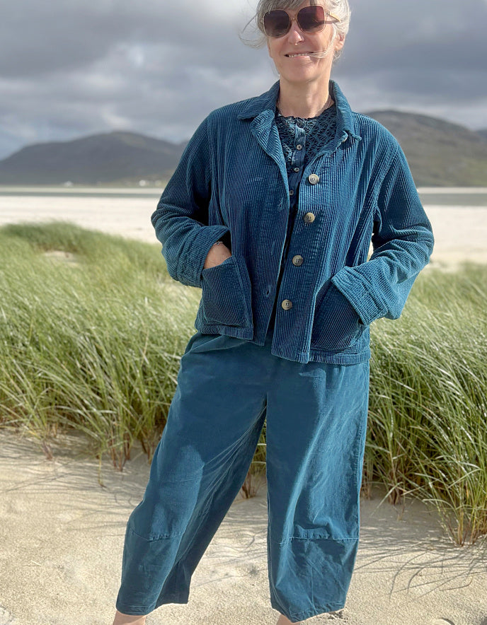 A woman wearing a matching blue corduroy jacket and wide-leg pants set stands on a sandy beach with dune grass and mountains in the background.