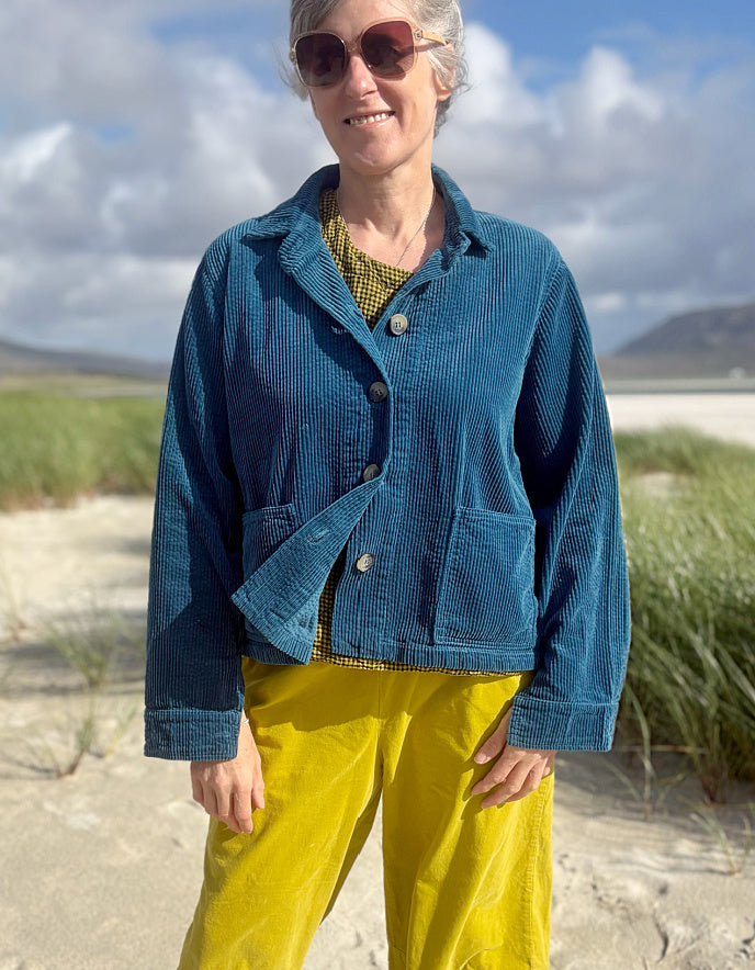 A woman wearing a textured blue corduroy jacket and bright yellow corduroy pants, standing outdoors on a sandy beach with dune grass and a cloudy sky in the background.