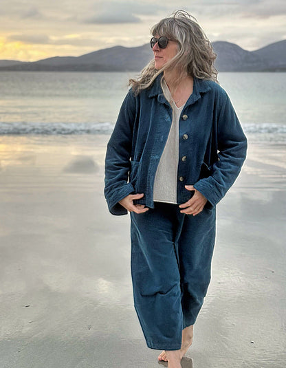 A woman stands barefoot on a beach at sunset wearing a dark blue corduroy jacket and matching wide-leg pants over a light gray top.