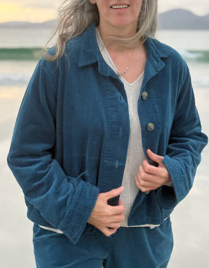 A woman wearing a blue corduroy jacket and matching pants over a white top, standing on a beach with water and distant hills in the background.