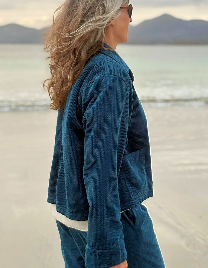 A woman standing on a beach wearing a textured blue corduroy jacket and matching blue corduroy pants, shown in profile with mountains and water in the background.