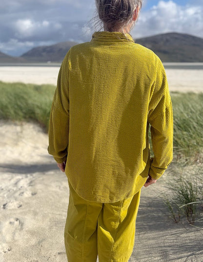 A person standing outdoors on a sandy path wearing a textured mustard yellow long-sleeve shirt and matching pants, with grass and distant hills under a cloudy sky in the background.
