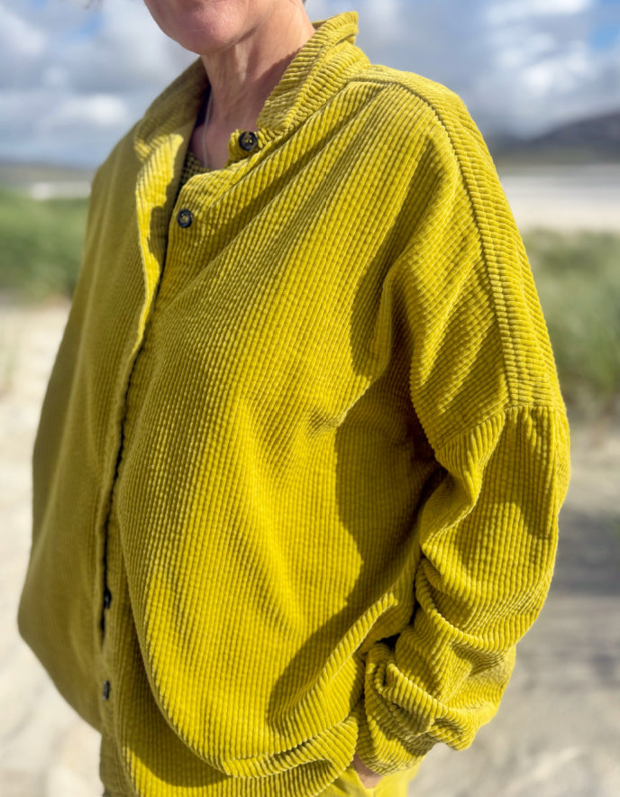A person wearing a textured mustard yellow corduroy button-up shirt jacket outdoors on a sandy beach with a cloudy sky in the background.