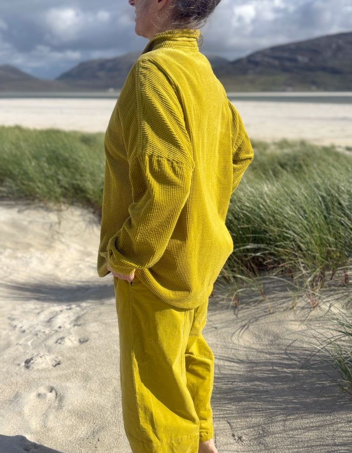 A person standing outdoors on a sandy beach path wearing a matching mustard yellow corduroy top and pants set, with grasses and mountains in the background.